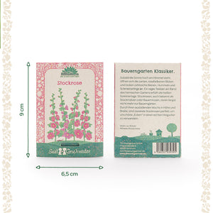 Dos paquetes de semillas de malvarrosa de Saatgeschwister, uno con una ilustración de las flores y otro con una descripción en alemán; dimensiones 9 cm de alto y 6,5 cm de ancho.