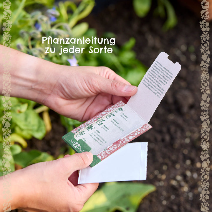 Manos sosteniendo un paquete de semillas abierto con instrucciones de siembra en el jardín, con plantas verdes y tierra en el fondo.