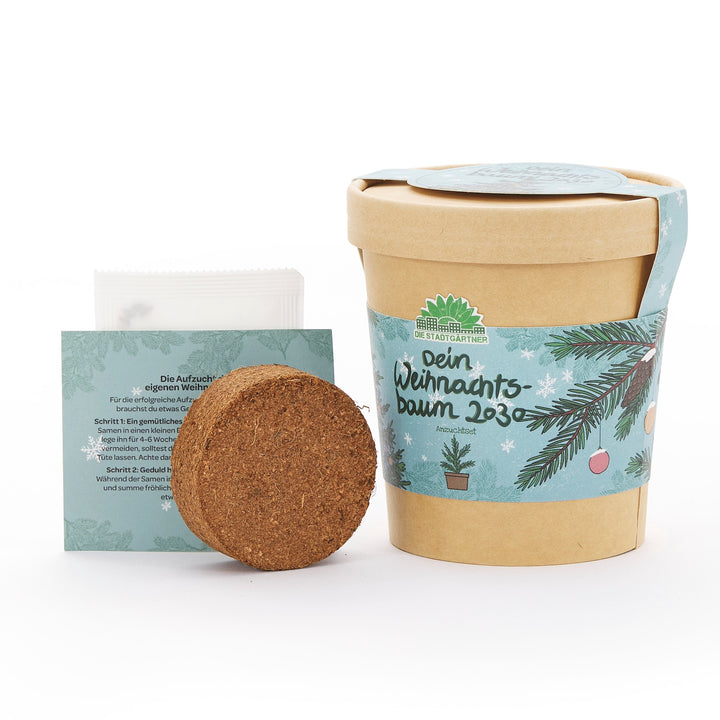 Un kit de cultivo para un árbol de Navidad, compuesto por una maceta marrón con una pancarta navideña, una pastilla de tierra para macetas e instrucciones en alemán.