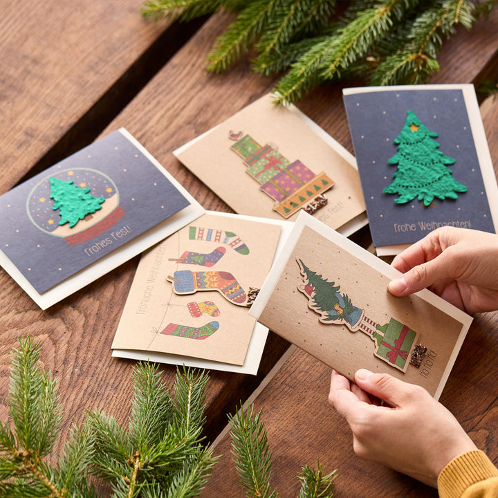 Varias tarjetas de felicitación navideñas con motivos coloridos y ramas de abeto sobre una mesa de madera, una de las tarjetas está sostenida por las manos.