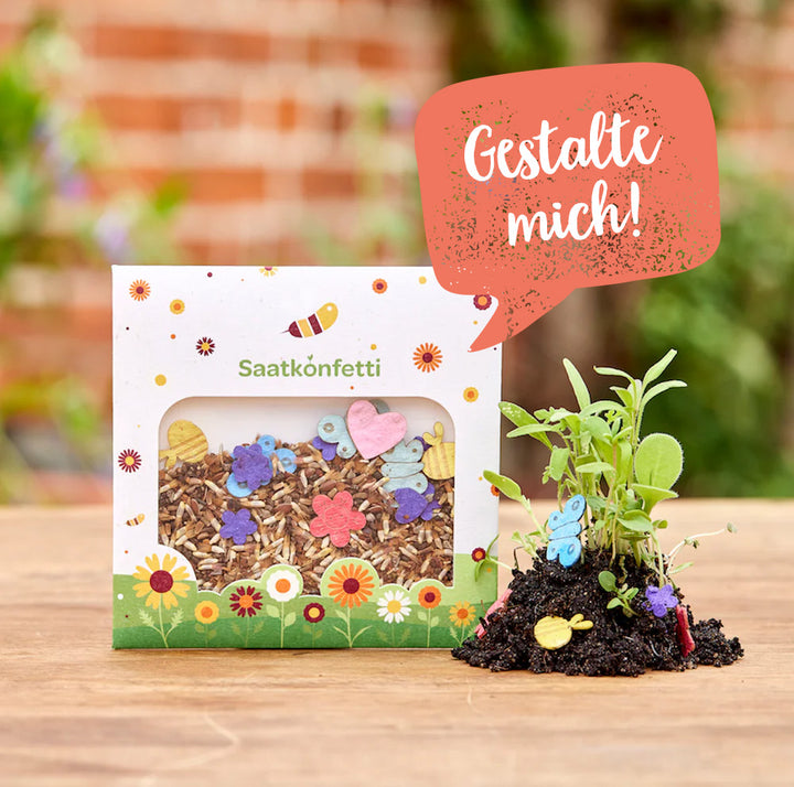 Paquete de confeti de semillas y pequeñas figuras de papel de colores, junto a plantas jóvenes, sobre una mesa de madera, con la naturaleza borrosa al fondo.