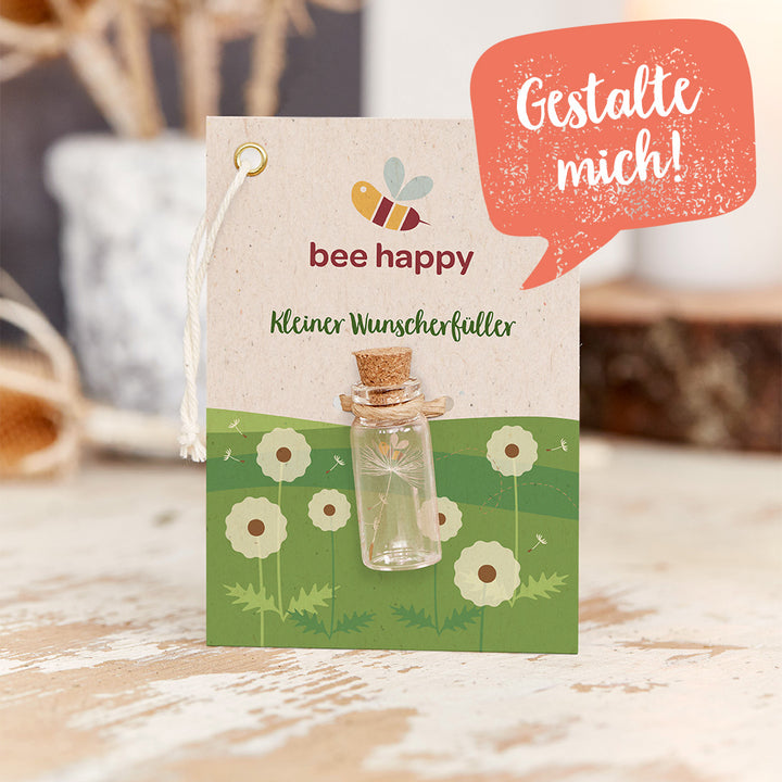 Tarjeta con la inscripción "abeja feliz" y "Pequeño concededor de deseos", una pequeña botella de vidrio adherida, un fondo con un prado dibujado y dientes de león, y un bocadillo de diálogo con el texto "¡Diséñame!".
