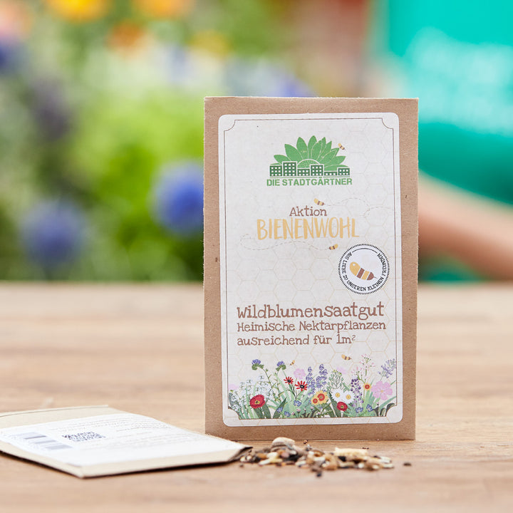 Bolsa de papel con semillas de flores silvestres “Aktion Bienenwohl” sobre una mesa de madera, flores de colores borrosas en el fondo.