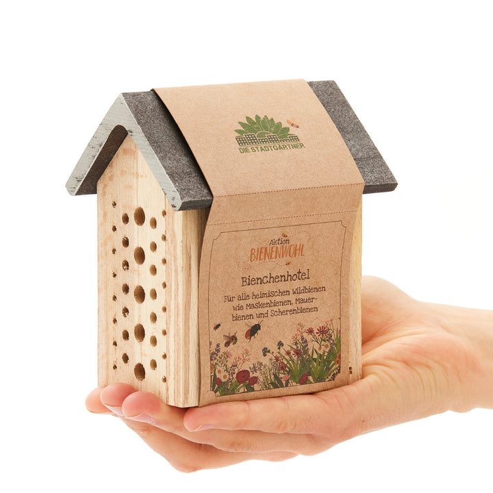 Una mano sostiene un pequeño hotel de madera para abejas con la inscripción "Aktion Bienenwohl Bienenhotel" en alemán.