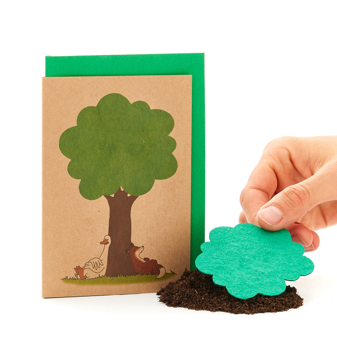 Una mano sostiene la copa de un árbol de papel verde delante de una tarjeta de felicitación con un tronco de árbol dibujado y animales sobre un fondo blanco.