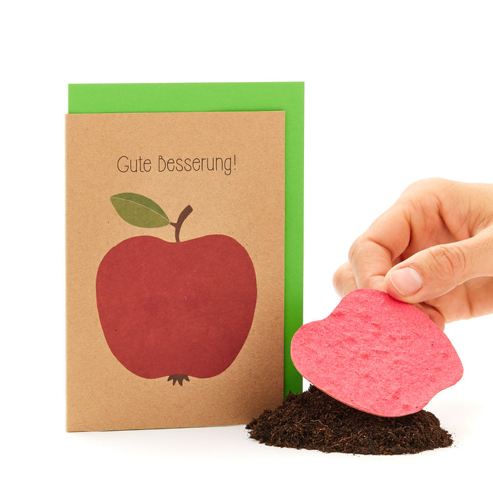 Una tarjeta de "que te mejores pronto" con una manzana encima junto a una mano que planta una tarjeta con semillas en forma de corazón en la tierra.