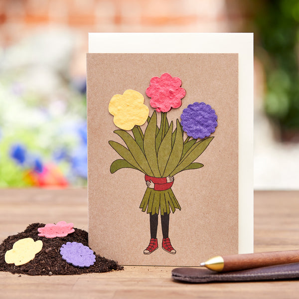 Tarjeta de felicitación hecha a mano con piernas dibujadas en zapatos rojos sosteniendo un ramo de flores de papel de colores sobre una mesa de madera con un bolígrafo al lado.