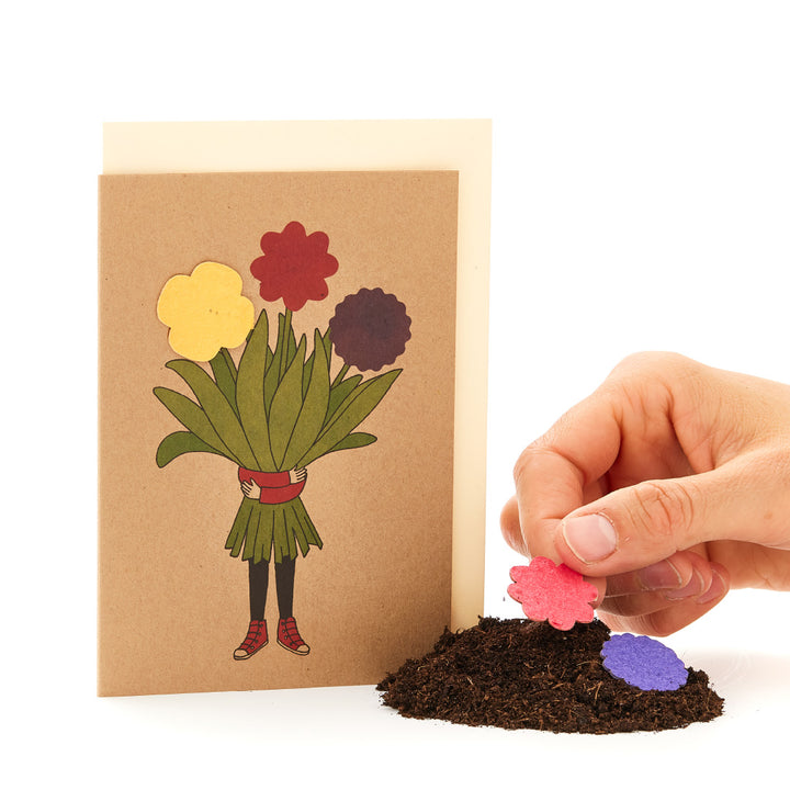 Mano colocando una flor de papel sobre un montón de tierra junto a una tarjeta de felicitación con un diseño de ramo.