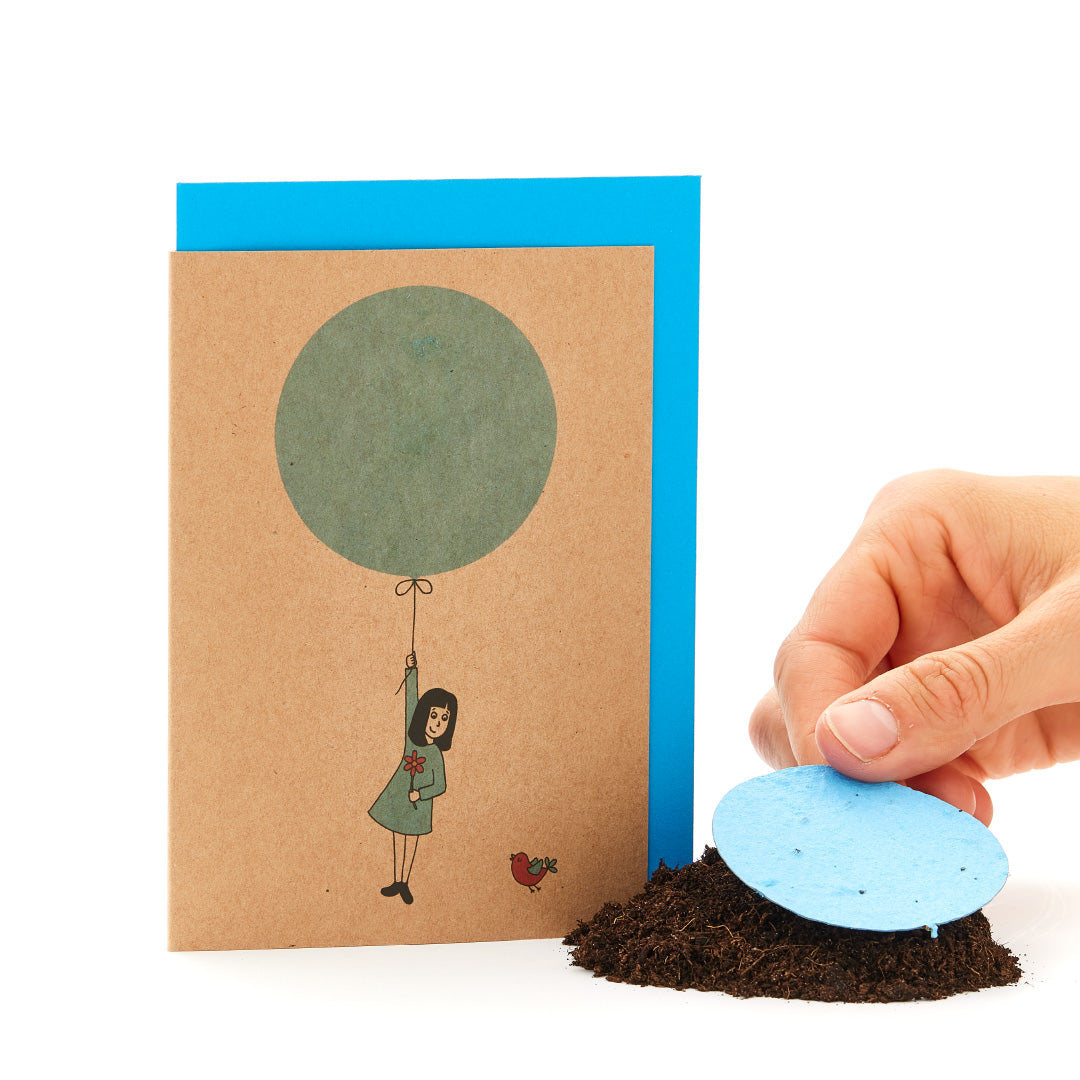 Una tarjeta de felicitación con una figura dibujada colgando de un globo y una mano colocando un papel con semillas redondo de color azul sobre el suelo.