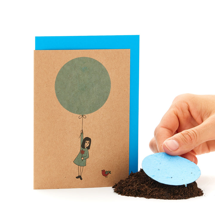 Una tarjeta de felicitación con una figura dibujada colgando de un globo y una mano colocando un papel con semillas redondo de color azul sobre el suelo.
