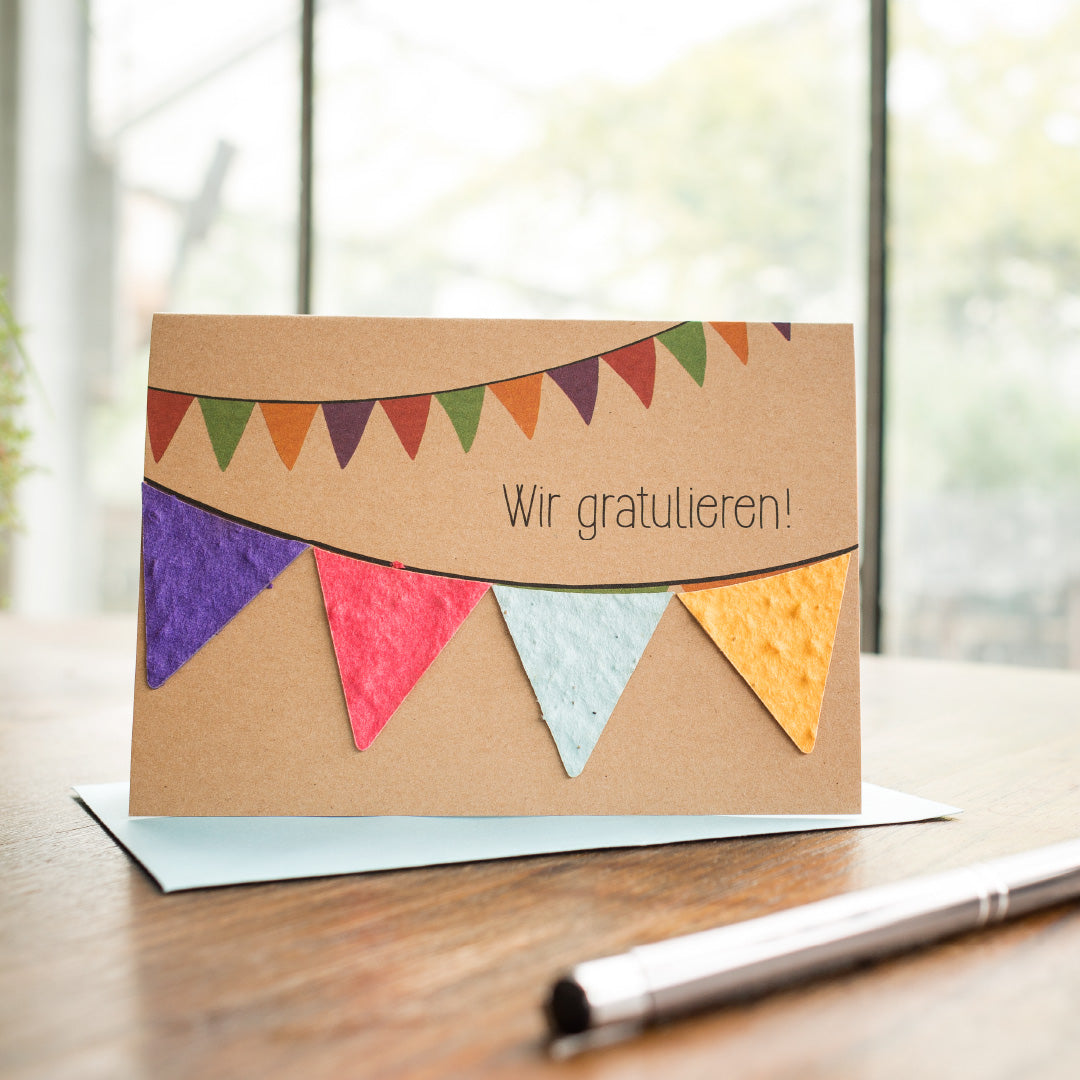 Tarjeta de felicitación con la inscripción "¡Felicitaciones!" y banderines de colores sobre una mesa de madera con un bolígrafo y un sobre.