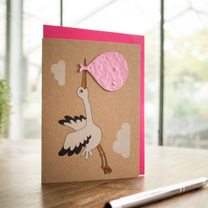 Tarjeta de felicitación con una cigüeña que lleva un paquete rosa sobre una mesa de madera con un bolígrafo al lado.