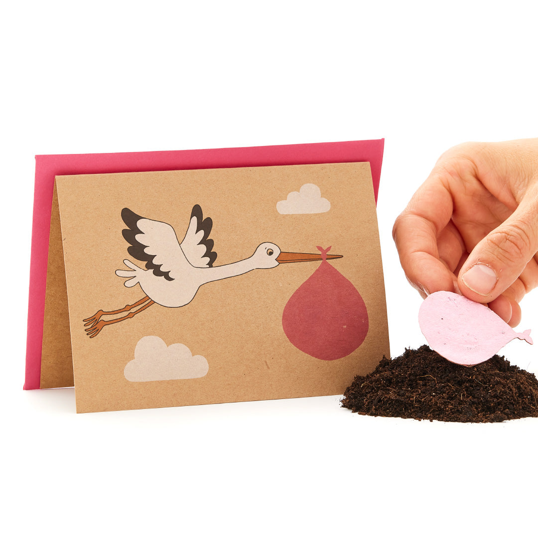 Cigüeña en una tarjeta de felicitación que lleva un paquete de bebés junto a una mano que sostiene un objeto rosa en forma de corazón sobre una pila de tierra.