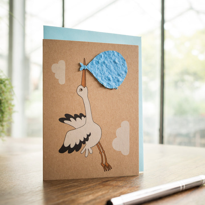 Tarjeta de felicitación hecha a mano con cigüeña llevando un bulto azul sobre una mesa con bolígrafo.