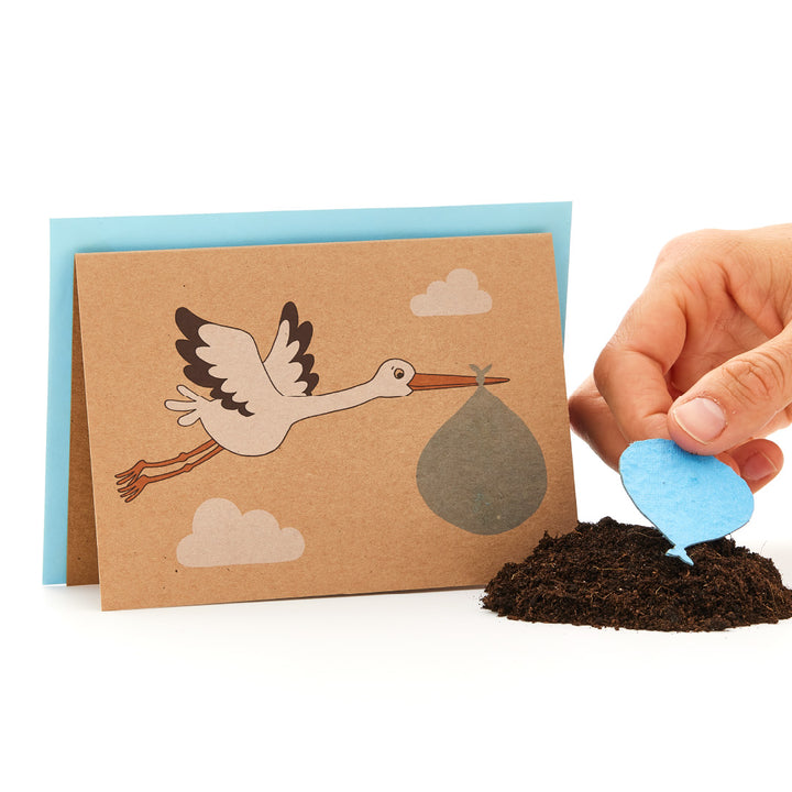 Tarjeta de felicitación con una cigüeña que lleva un paquete de bebés junto a una mano que planta una semilla azul en forma de corazón en el suelo.