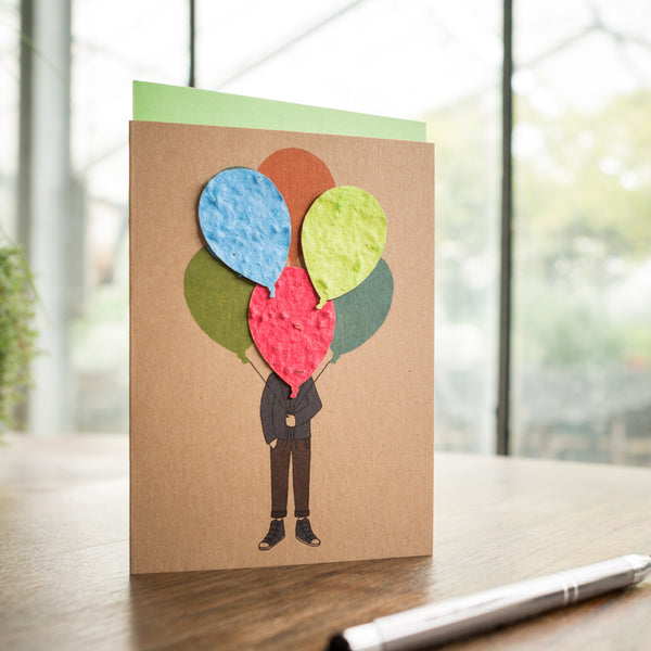 Tarjeta de felicitación con la ilustración de una persona cuya cabeza está hecha de globos de papel de colores, sobre una mesa junto a un bolígrafo.