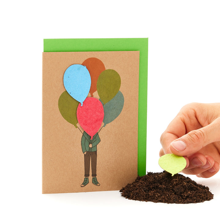Tarjeta de felicitación con una figura ilustrada que sostiene globos junto a un montón de tierra de la que crece un globo real.