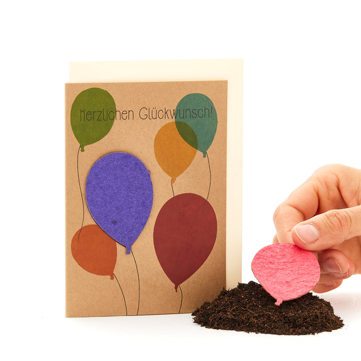 Tarjeta de felicitación con globos de colores y las palabras "Felicitaciones" junto a un montón de tierra y una mano plantando un globo rosa.