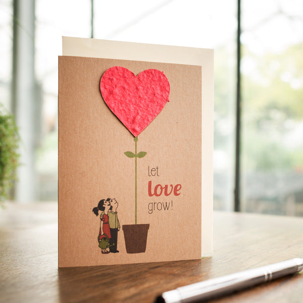 Tarjeta de felicitación con una pareja besándose junto a una maceta con una planta creciendo en ella con un corazón rojo, con el texto "¡Deja que el amor crezca!"