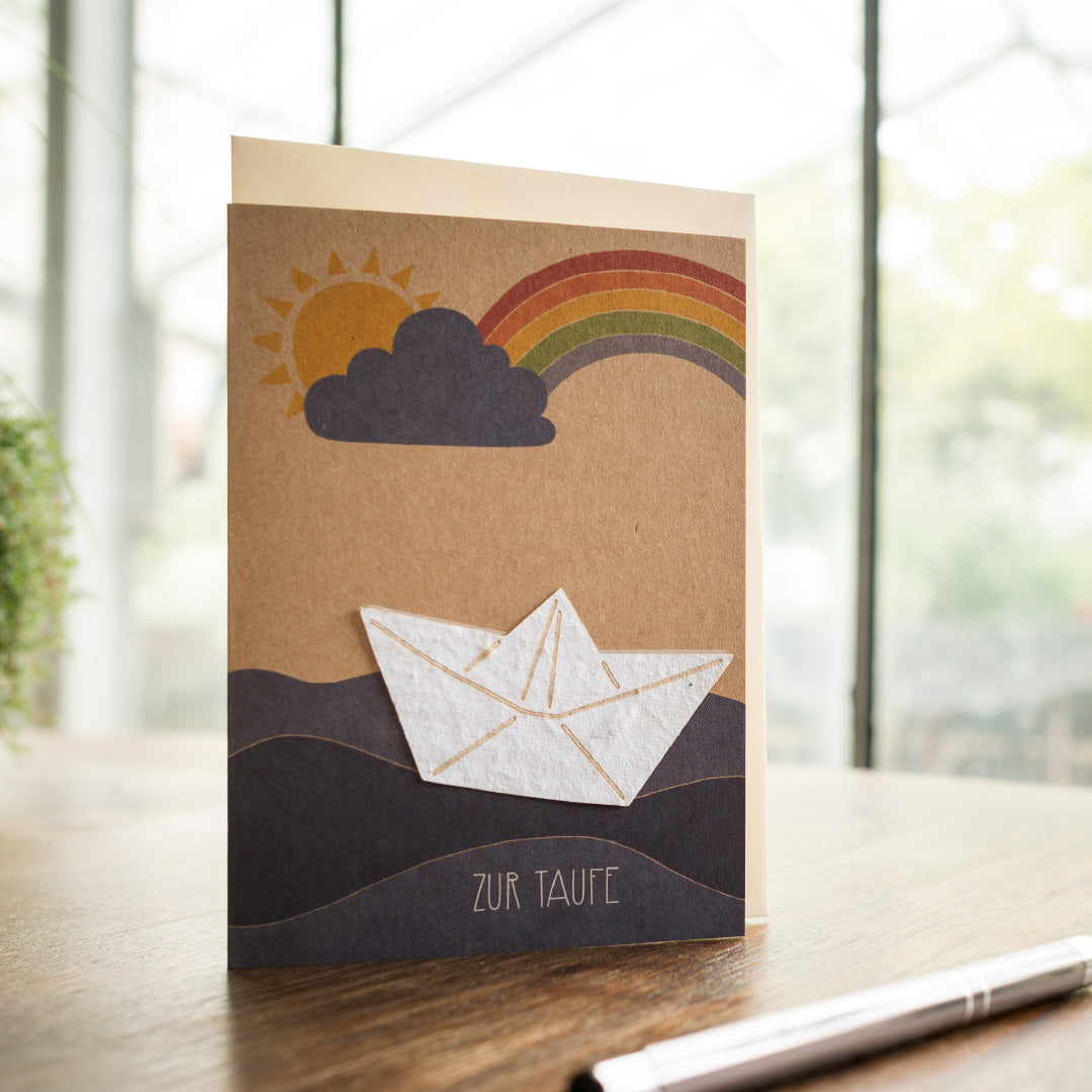 Tarjeta de bautismo con barquito de papel y arcoiris sobre mesa de madera.