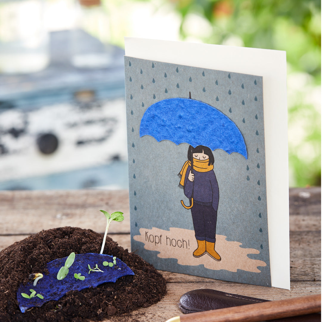 Tarjeta de felicitación con el dibujo de una persona bajo un paraguas azul y la inscripción "¡Ánimo!", junto a una planta joven en el suelo.