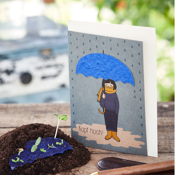 Tarjeta de felicitación con el dibujo de una persona bajo un paraguas azul y la inscripción "¡Ánimo!", junto a una planta joven en el suelo.