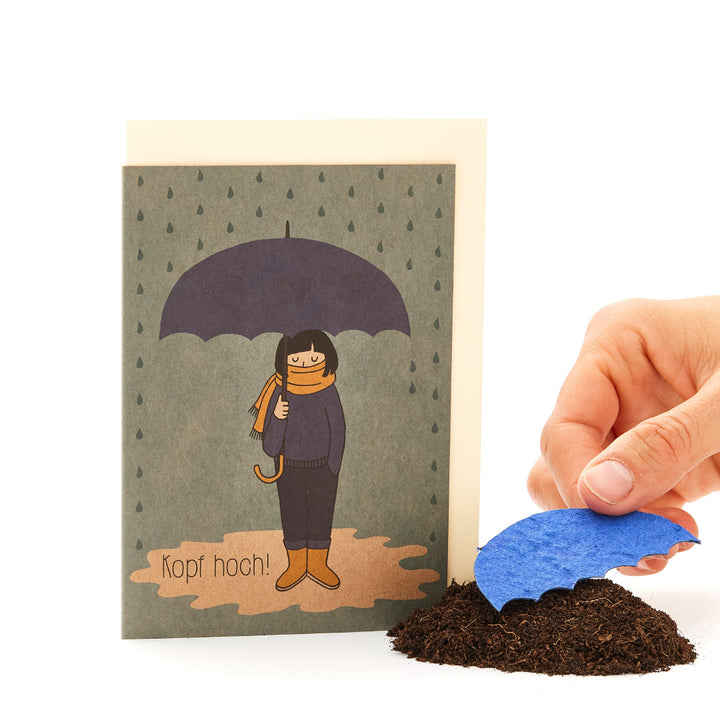 Mano colocando una ola de papel azul sobre un montón de tierra delante de una tarjeta de felicitación con una figura dibujada bajo un paraguas y el texto "¡Ánimo!"