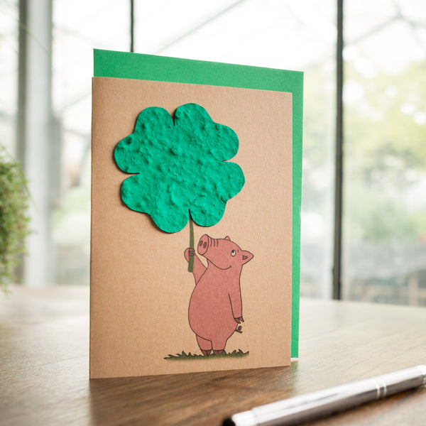 Tarjeta de felicitación hecha a mano con un cerdito dibujado sosteniendo un árbol de papel verde sobre una mesa de madera con un bolígrafo.