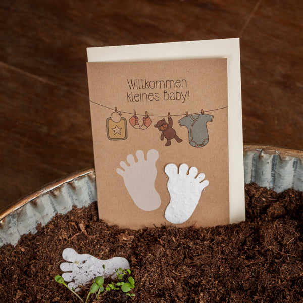 Tarjeta de baby shower con la inscripción "¡Bienvenido bebé!" e ilustraciones de pies de bebé, una percha con un pelele y juguetes, sobre un fondo de madera sucia.