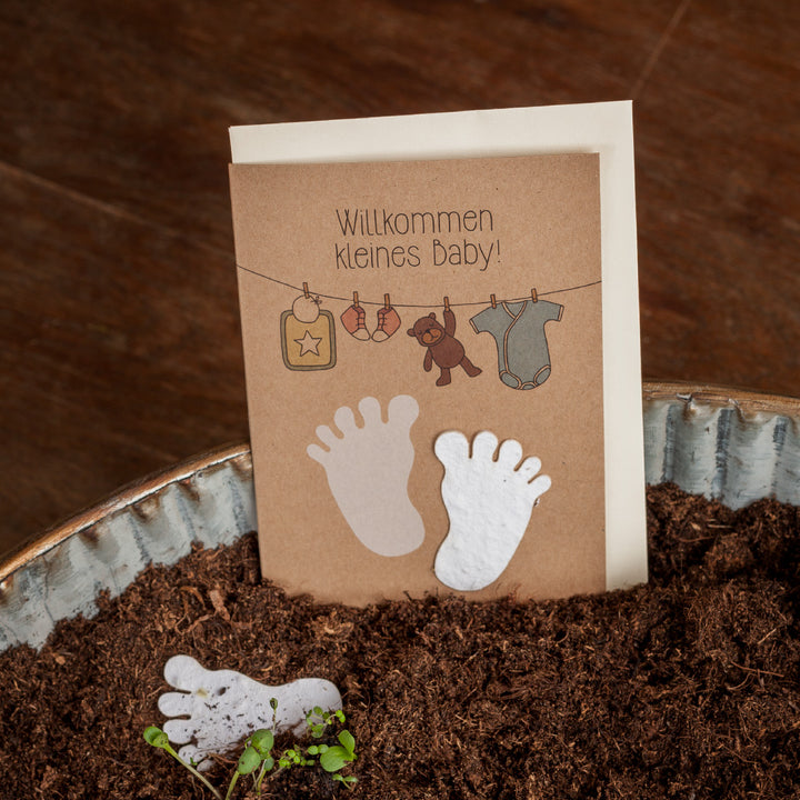Tarjeta de baby shower con la inscripción "¡Bienvenido bebé!" e ilustraciones de pies de bebé, una percha con un pelele y juguetes, sobre un fondo de madera sucia.