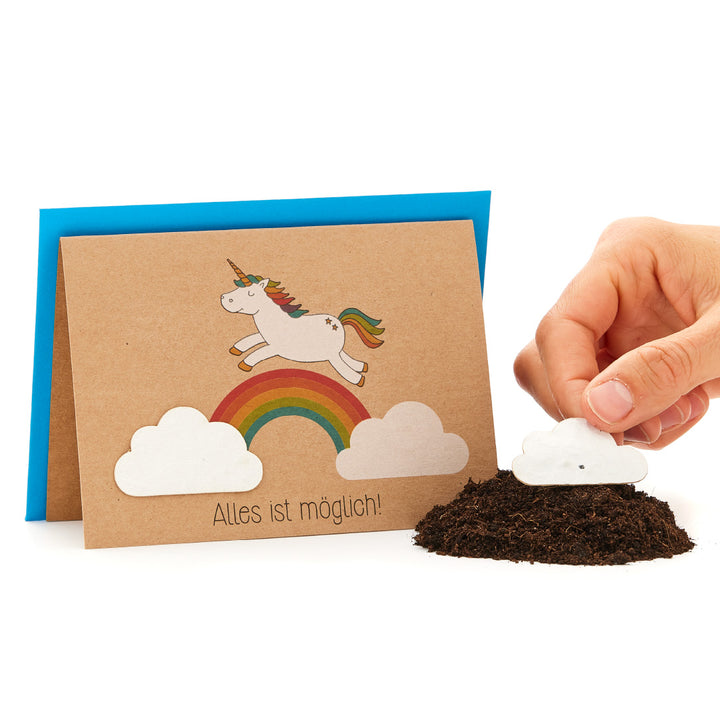 Una mano planta una semilla de papel en forma de nube en la tierra, junto a una tarjeta de felicitación con un unicornio sobre un arcoíris y el dicho "¡Todo es posible!".