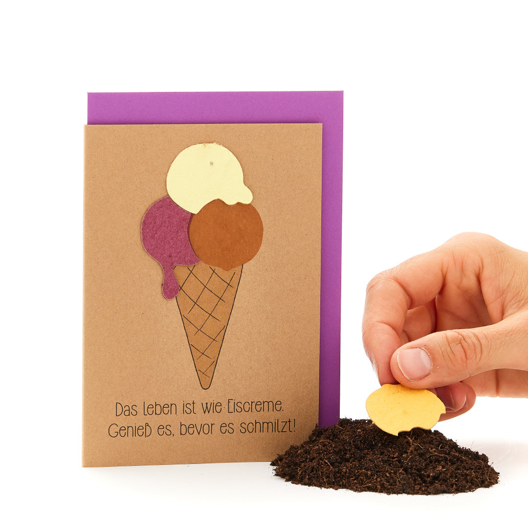 Una mano clava una galleta con forma de corazón en la tierra, junto a una tarjeta de felicitación en forma de cono de helado con el lema «La vida es como el helado. ¡Disfrútalo antes de que se derrita!».