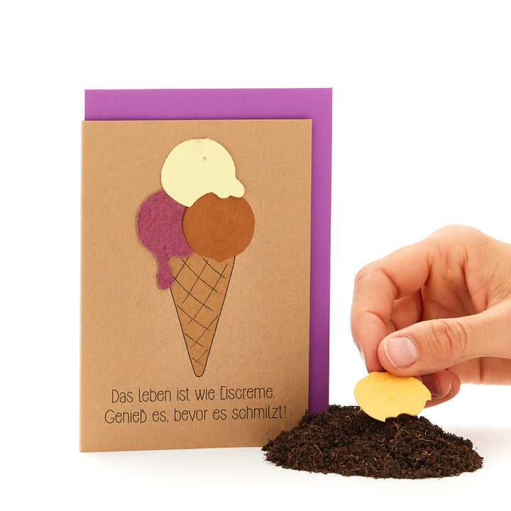 Una mano clava una galleta con forma de corazón en la tierra, junto a una tarjeta de felicitación en forma de cono de helado con el lema «La vida es como el helado. ¡Disfrútalo antes de que se derrita!».