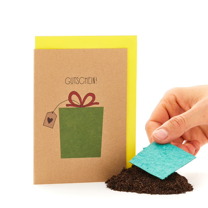 Mano sosteniendo una tarjeta de felicitación con la inscripción "VOUCHER" y un regalo dibujado, junto a un montón de tierra con un trozo de cartón azul.