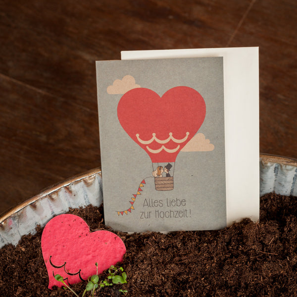 Tarjeta de felicitación de boda que presenta un globo aerostático en forma de corazón y la inscripción "¡Feliz boda!", colocado junto a un objeto en forma de corazón y tierra para macetas.