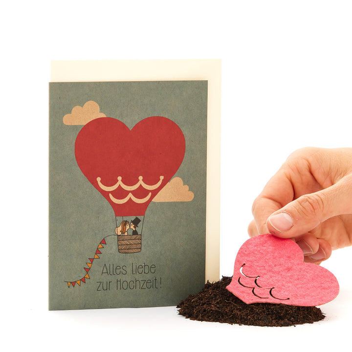 Tarjeta de felicitación de boda con un globo aerostático en forma de corazón y una mano plantando un corazón de papel en el suelo.