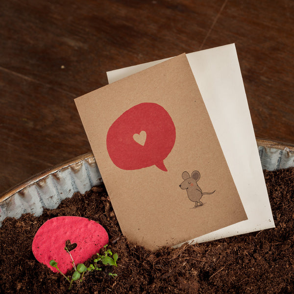 Una tarjeta de felicitación con un ratón dibujado y un bocadillo de diálogo rojo con un corazón, al lado un sobre blanco y un corazón de papel rojo en la tierra.
