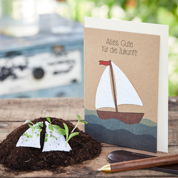 Tarjeta de felicitación con un velero y la inscripción "¡Todo lo mejor para el futuro!", junto a él tierra con plántulas y un bolígrafo sobre una mesa de madera.