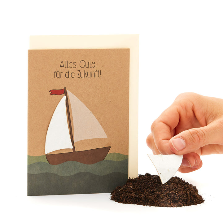 Tarjeta de felicitación con motivo de velero y la inscripción “¡Todo lo mejor para el futuro!”, junto a ella una mano clavando un trozo de papel con forma de vela en el suelo.