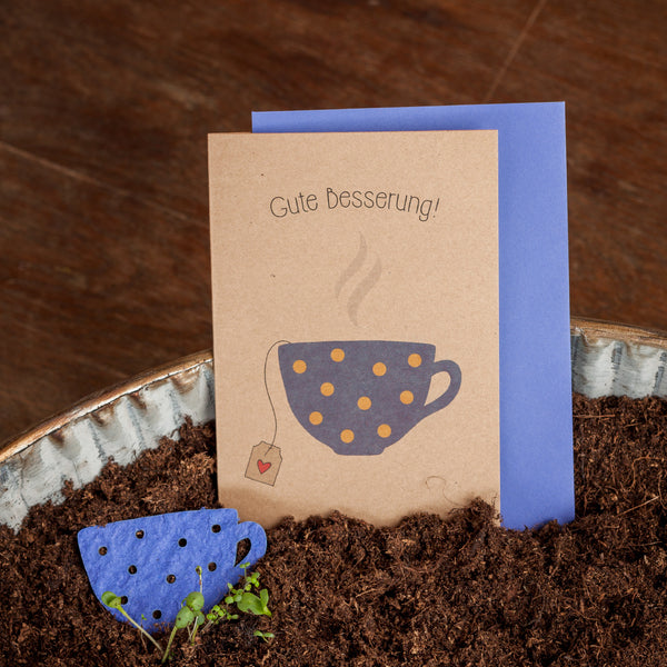Tarjeta de pronta recuperación con las palabras "¡Mejórate pronto!" y una taza ilustrada en el frente, junto a un adorno para vaso de papel azul en color tierra.