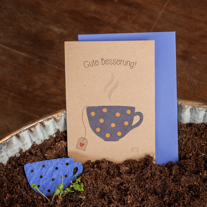 Tarjeta de pronta recuperación con las palabras "¡Mejórate pronto!" y una taza ilustrada en el frente, junto a un adorno para vaso de papel azul en color tierra.