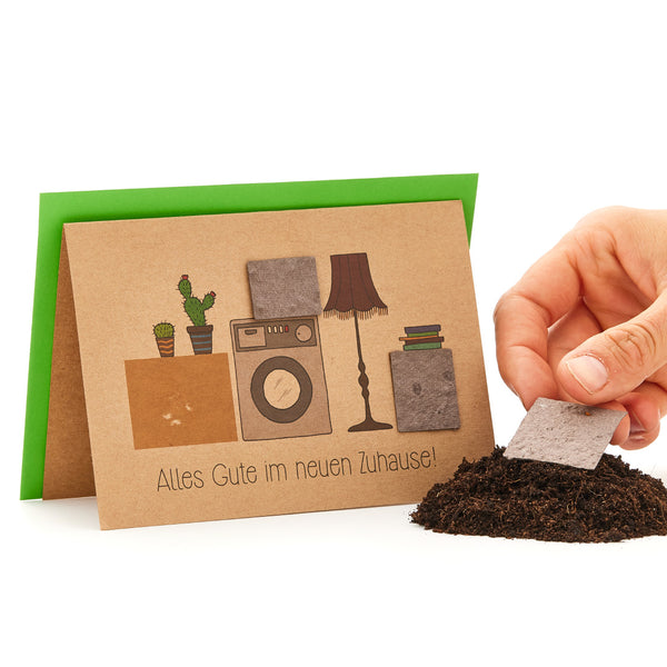 Tarjeta de felicitación con la inscripción "Todo lo mejor en tu nuevo hogar" y elementos del hogar dibujados, junto a un montón de tierra con un paño con semillas de plantas.