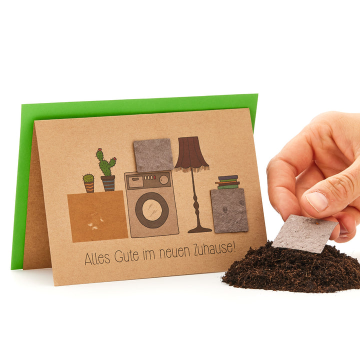 Tarjeta de felicitación con la inscripción "Todo lo mejor en tu nuevo hogar" y elementos del hogar dibujados, junto a un montón de tierra con un paño con semillas de plantas.
