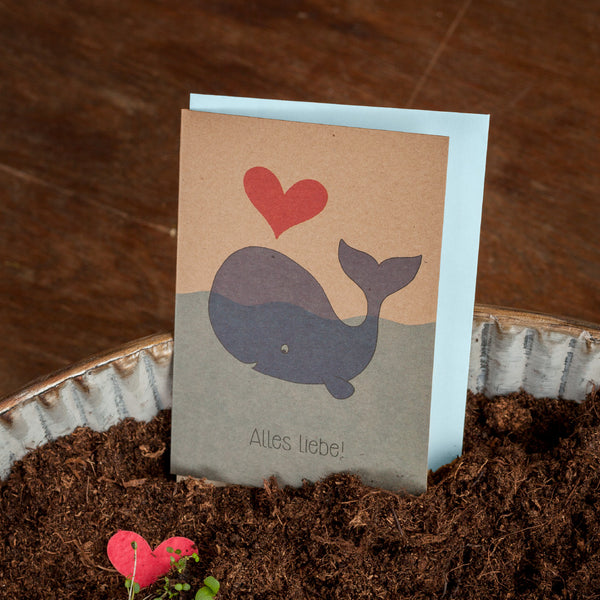 Tarjeta de felicitación con una ballena dibujada, un corazón rojo y el texto "¡Todo lo mejor!" de pie sobre la tierra.