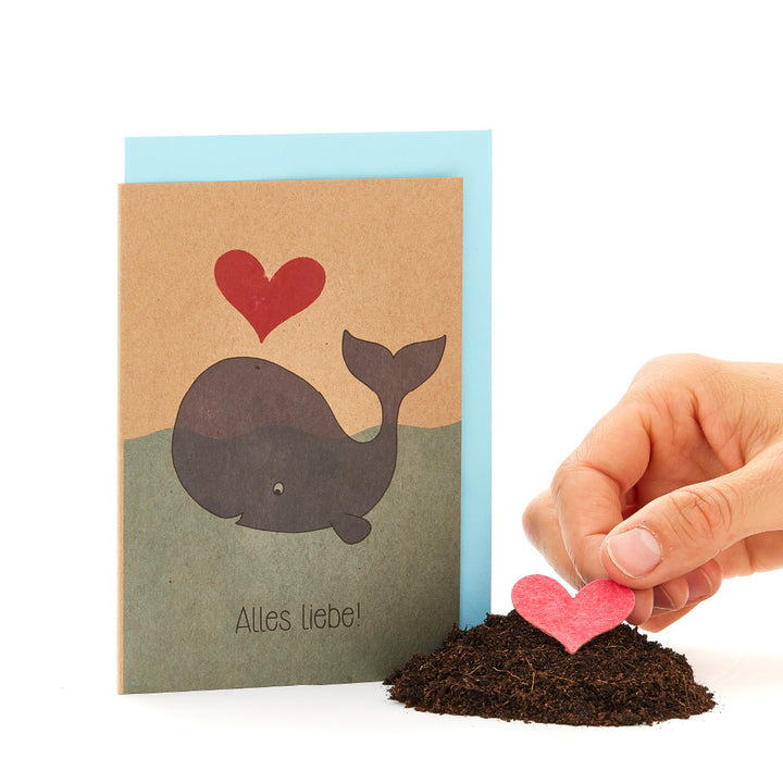 Tarjeta de felicitación con una ballena impresa y un corazón rojo, al lado una mano plantando un corazón rojo en el suelo.