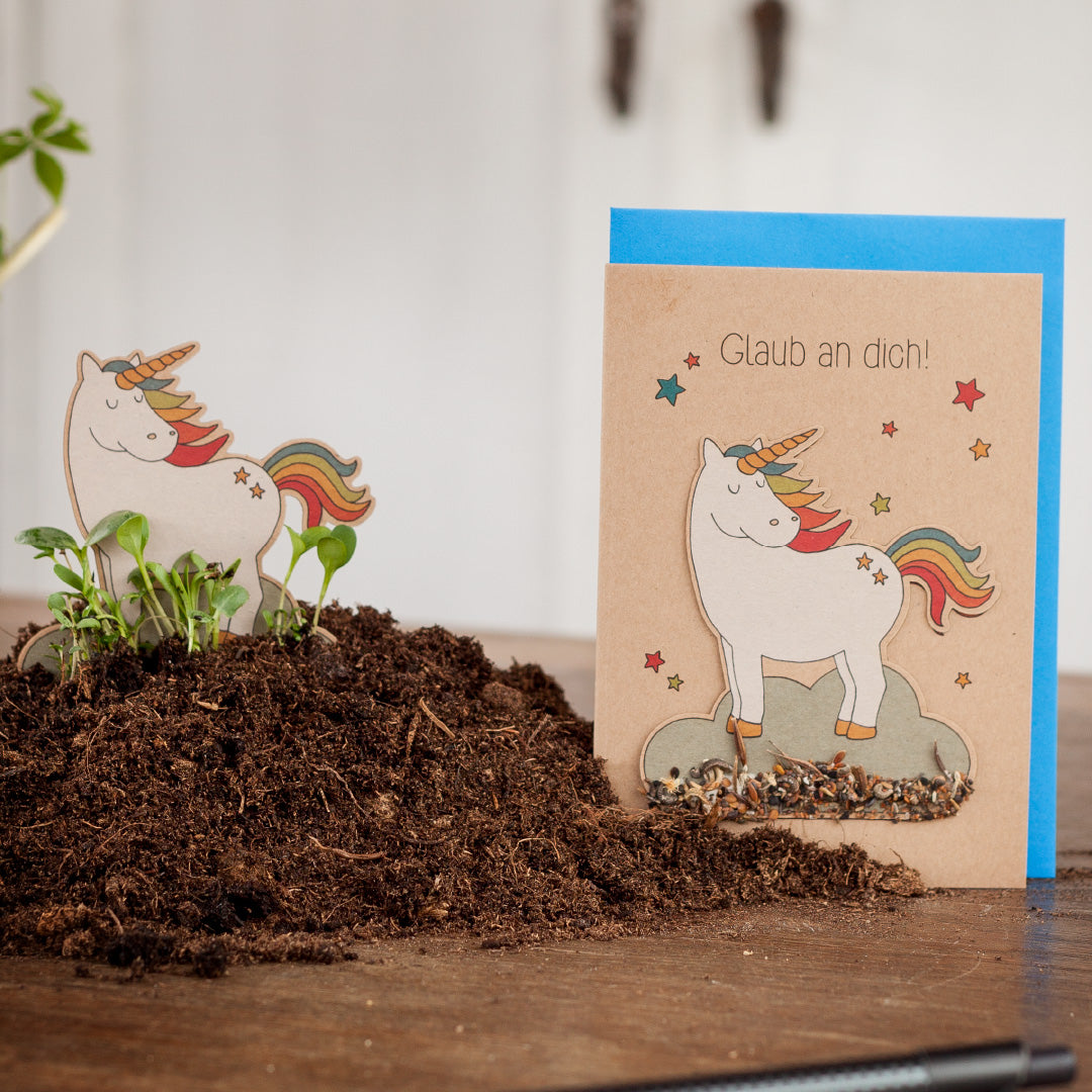 Una tarjeta de felicitación con un unicornio y el texto "¡Cree en ti mismo!" junto a un pequeño recorte de unicornio rodeado de tierra.