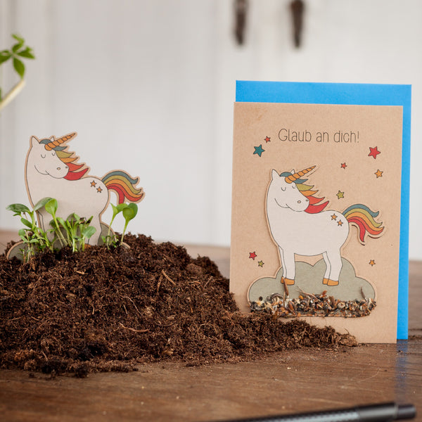 Una tarjeta de felicitación con un unicornio y el texto "¡Cree en ti mismo!" junto a un pequeño recorte de unicornio rodeado de tierra.