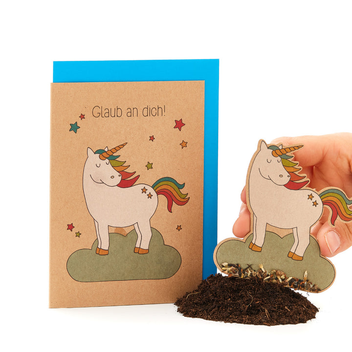 Tarjeta de felicitación con un unicornio y un arcoíris, el texto "¡Cree en ti mismo!" y una mano clavando un papel de unicornio a juego en la tierra.