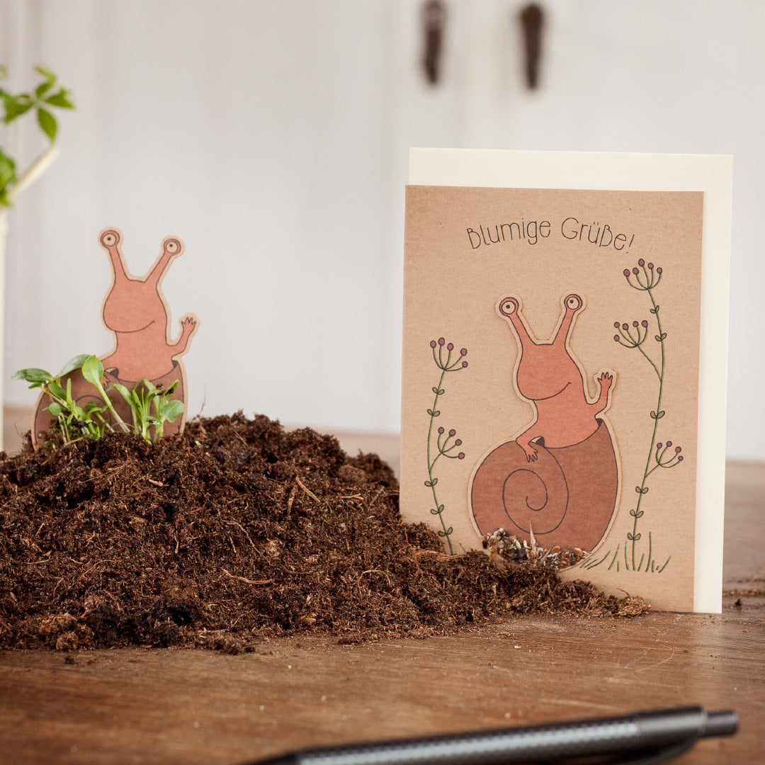 Tarjeta de felicitación con motivo de caracol y la inscripción "saludos florales" junto a un montón de tierra sobre una mesa de madera.