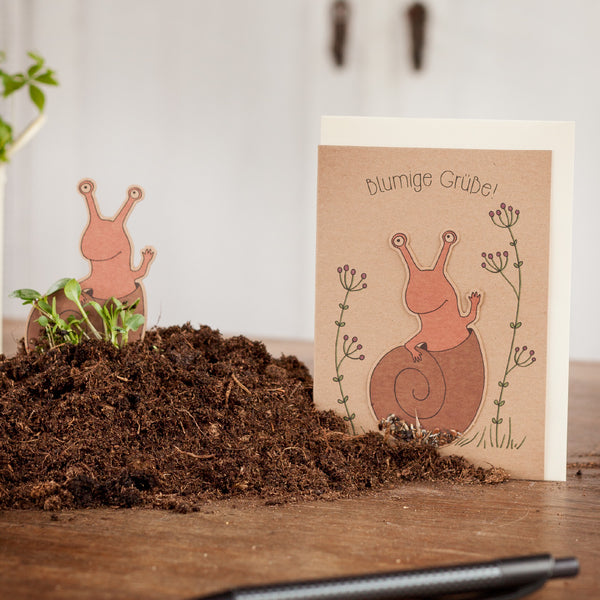 Tarjeta de felicitación con motivo de caracol y la inscripción "saludos florales" junto a un montón de tierra sobre una mesa de madera.